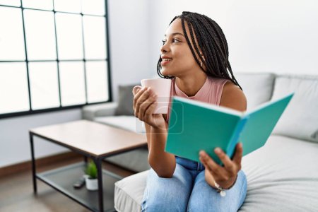 Foto de Joven mujer afroamericana leyendo libro y tomando café en casa - Imagen libre de derechos