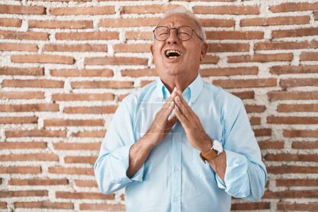 Téléchargez les photos : Homme âgé avec les cheveux gris debout sur le mur de briques mendier et prier avec les mains ainsi que l'expression de l'espoir sur le visage très émotionnel et inquiet. mendier. - en image libre de droit
