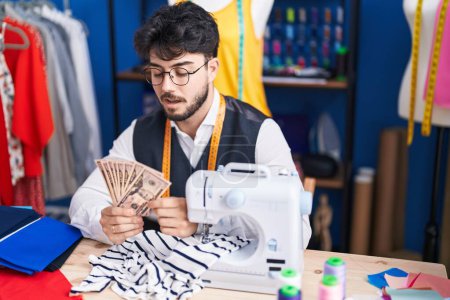 Téléchargez les photos : Young hispanic man tailor holding dollars at sewing studio - en image libre de droit