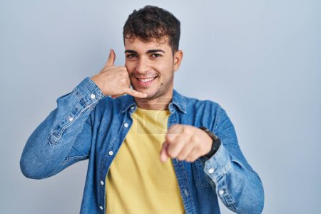 Téléchargez les photos : Young hispanic man standing over blue background smiling doing talking on the telephone gesture and pointing to you. call me. - en image libre de droit