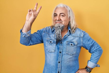 Photo for Middle age man with grey hair standing over yellow background smiling looking to the camera showing fingers doing victory sign. number two. - Royalty Free Image