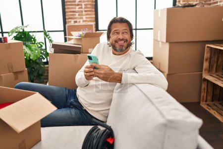 Foto de Hombre de mediana edad con teléfono inteligente sentado en el sofá en el nuevo hogar - Imagen libre de derechos