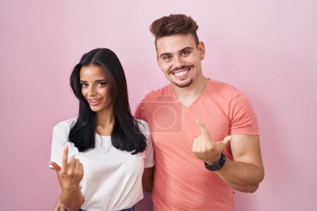Foto de Joven pareja hispana de pie sobre fondo rosa haciendo señas ven aquí gesto con la mano invitando a la bienvenida feliz y sonriente - Imagen libre de derechos