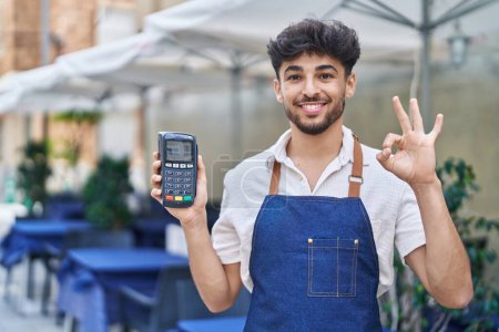 Téléchargez les photos : Arabe homme avec barbe portant tablier de serveur à la terrasse du restaurant tenant dataphone faire ok signe avec les doigts, sourire geste amical excellent symbole - en image libre de droit