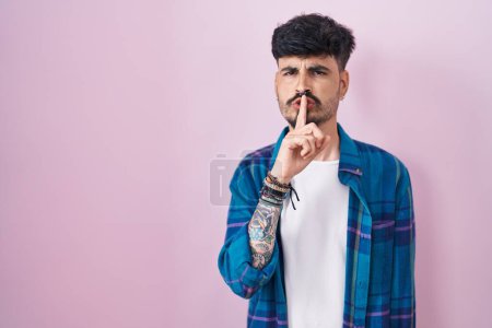 Foto de Young hispanic man with beard standing over pink background asking to be quiet with finger on lips. silence and secret concept. - Imagen libre de derechos