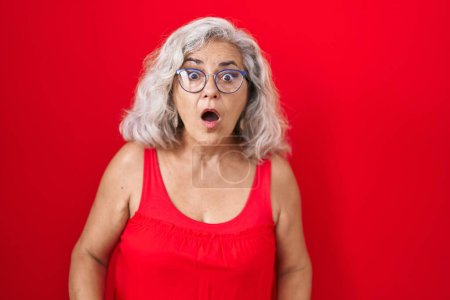 Téléchargez les photos : Middle age woman with grey hair standing over red background afraid and shocked with surprise and amazed expression, fear and excited face. - en image libre de droit
