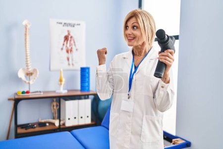 Téléchargez les photos : Middle age blonde woman holding therapy massage gun at physiotherapy center pointing thumb up to the side smiling happy with open mouth - en image libre de droit
