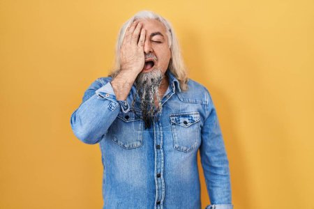 Foto de Middle age man with grey hair standing over yellow background yawning tired covering half face, eye and mouth with hand. face hurts in pain. - Imagen libre de derechos