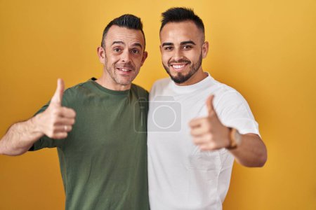 Foto de Homosexual couple standing over yellow background approving doing positive gesture with hand, thumbs up smiling and happy for success. winner gesture. - Imagen libre de derechos