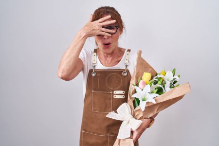 Téléchargez les photos : Femme d'âge moyen tenant bouquet de fleurs blanches regardant dans le choc couvrant le visage et les yeux avec la main, regardant à travers les doigts avec une expression embarrassée. - en image libre de droit