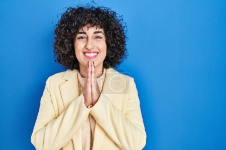 Téléchargez les photos : Young brunette woman with curly hair standing over blue background praying with hands together asking for forgiveness smiling confident. - en image libre de droit