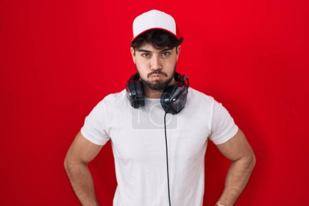 Foto de Hispanic man with beard wearing gamer hat and headphones puffing cheeks with funny face. mouth inflated with air, crazy expression. - Imagen libre de derechos