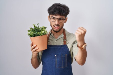 Téléchargez les photos : Arab man with beard holding green plant pot angry and mad raising fist frustrated and furious while shouting with anger. rage and aggressive concept. - en image libre de droit