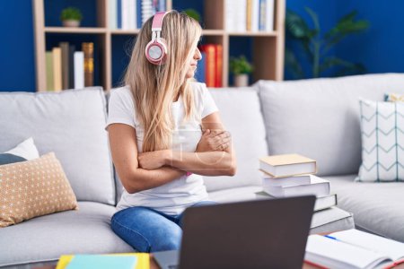 Téléchargez les photos : Jeune femme blonde étudiant à l'aide d'un ordinateur portable à la maison regardant vers le côté avec les bras croisés convaincus et confiants - en image libre de droit