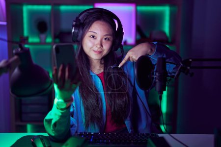 Foto de Joven mujer asiática jugando videojuegos con smartphone mirando confiado con sonrisa en la cara, apuntándose con los dedos orgullosos y felices. - Imagen libre de derechos