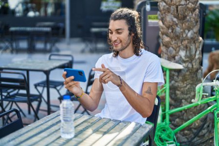 Photo for Young hispanic man watching video on smartphone sitting on table at coffee shop terrace - Royalty Free Image