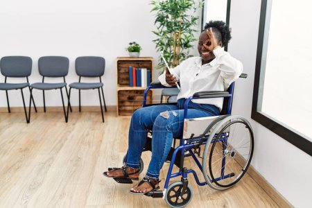Téléchargez les photos : Jeune femme noire assise en fauteuil roulant dans la salle d'attente faisant un geste correct avec la main souriante, les yeux regardant à travers les doigts avec un visage heureux. - en image libre de droit