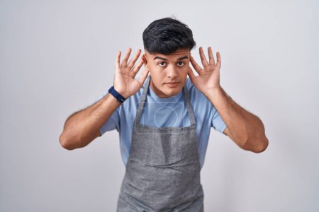 Téléchargez les photos : Jeune homme hispanique portant tablier sur fond blanc essayant d'entendre les deux mains sur le geste de l'oreille, curieux de commérages. problème auditif, sourd - en image libre de droit
