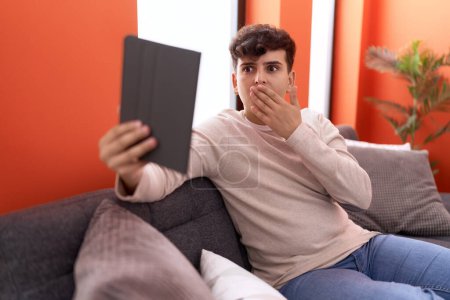 Téléchargez les photos : Young non binary man using touchpad sitting on the sofa covering mouth with hand, shocked and afraid for mistake. surprised expression - en image libre de droit
