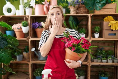 Foto de Mujer joven caucásica que trabaja en floristería sosteniendo la planta cubriendo la boca con la mano, conmocionada y temerosa de equivocarse. expresión sorprendida - Imagen libre de derechos