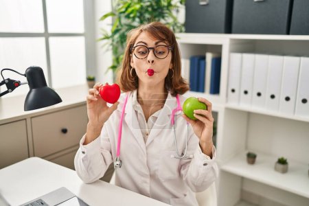 Téléchargez les photos : Young doctor woman holding heart and green apple making fish face with mouth and squinting eyes, crazy and comical. - en image libre de droit