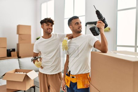 Téléchargez les photos : Deux hommes hispaniques couple souriant maison réparatrice confiante à la nouvelle maison - en image libre de droit