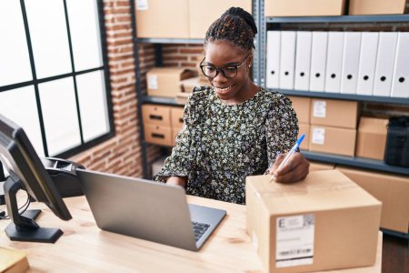 Photo for African american woman ecommerce business worker using laptop writing on package at office - Royalty Free Image