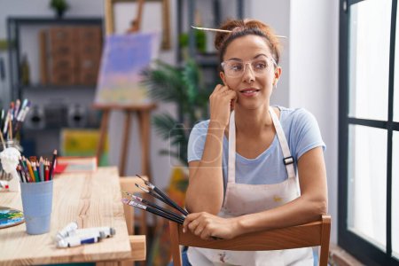 Téléchargez les photos : Young woman artist sitting on chair holding paintbrush at art studio - en image libre de droit