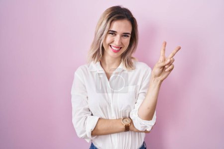 Photo for Young beautiful woman standing over pink background smiling with happy face winking at the camera doing victory sign with fingers. number two. - Royalty Free Image
