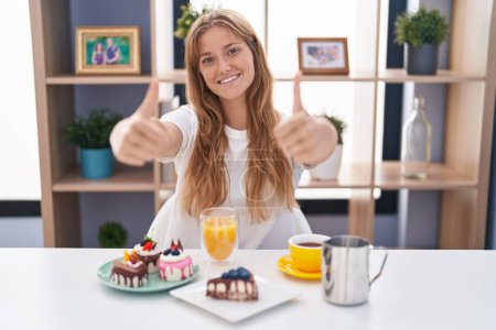 Foto de Joven mujer caucásica comiendo pasteles t para el desayuno aprobando hacer gesto positivo con la mano, pulgares arriba sonriendo y feliz por el éxito. gesto ganador. - Imagen libre de derechos