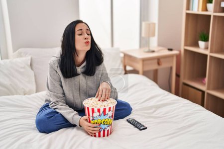 Foto de Hispanic woman eating popcorn watching a movie on the bed looking at the camera blowing a kiss being lovely and sexy. love expression. - Imagen libre de derechos