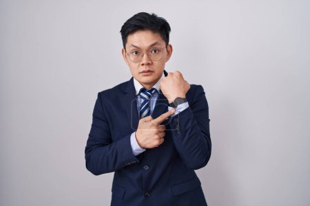 Téléchargez les photos : Jeune homme asiatique portant costume d'affaires et cravate à la hâte pointant pour regarder le temps, l'impatience, en regardant la caméra avec une expression détendue - en image libre de droit