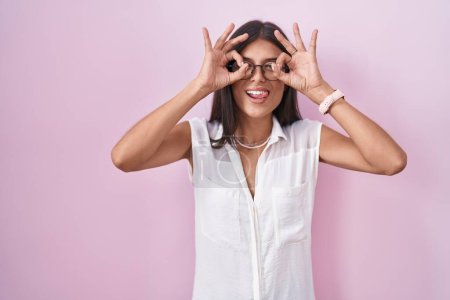 Foto de Brunette young woman standing over pink background wearing glasses doing ok gesture like binoculars sticking tongue out, eyes looking through fingers. crazy expression. - Imagen libre de derechos