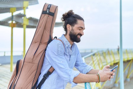 Téléchargez les photos : Jeune musicien hispanique utilisant un smartphone tenant un étui de guitare au bord de la mer - en image libre de droit