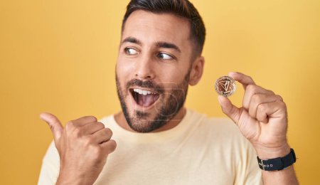 Photo for Handsome hispanic man holding tron cryptocurrency coin pointing thumb up to the side smiling happy with open mouth - Royalty Free Image