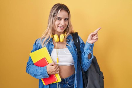 Téléchargez les photos : Jeune femme blonde portant un sac à dos étudiant et tenant des livres avec un grand sourire sur le visage, pointant du doigt vers le côté en regardant la caméra. - en image libre de droit