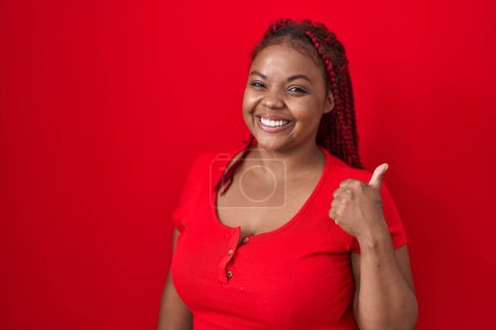 Téléchargez les photos : Femme afro-américaine avec des cheveux tressés debout sur fond rouge faisant des pouces heureux geste avec la main. approuvant l'expression regardant la caméra montrant le succès. - en image libre de droit