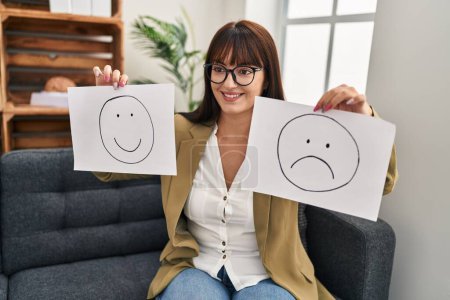 Foto de Joven hermosa mujer hispana psicóloga sosteniendo tristeza y sonriente emoji documentos en la clínica de psicología - Imagen libre de derechos