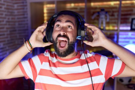 Photo for Hispanic man with beard playing video games wearing headphones angry and mad screaming frustrated and furious, shouting with anger looking up. - Royalty Free Image
