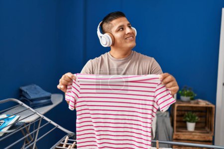 Téléchargez les photos : Jeune homme latin écoutant de la musique accrochant des vêtements sur la corde à linge à la buanderie - en image libre de droit