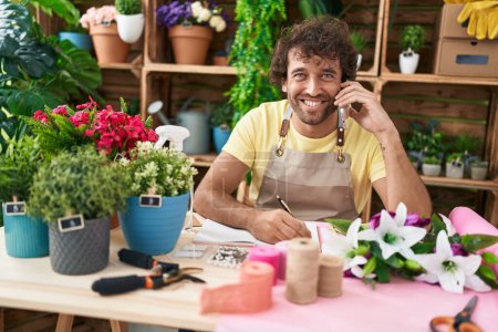 Photo for Young hispanic man florist talking on smartphone writing on notebook at flower shop - Royalty Free Image