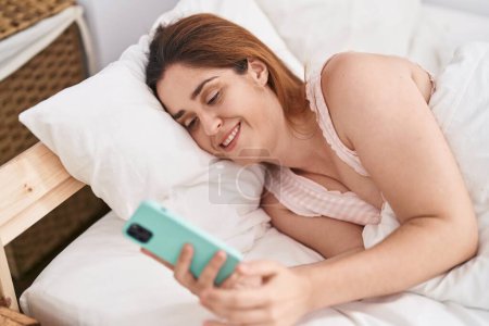 Téléchargez les photos : Jeune femme utilisant smartphone couché sur le lit dans la chambre - en image libre de droit