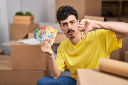 Téléchargez les photos : Homme hispanique dans une nouvelle maison tenant des billets en francs suisses avec un visage en colère, signe négatif déplaisant avec les pouces baissés, concept de rejet - en image libre de droit