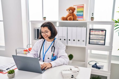Téléchargez les photos : Jeune femme portant l'uniforme de médecin en utilisant un ordinateur portable travaillant à la clinique - en image libre de droit