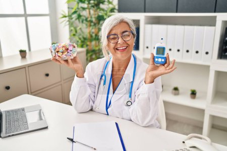 Photo for Middle age woman with grey hair wearing doctor uniform holding glucose monitor winking looking at the camera with sexy expression, cheerful and happy face. - Royalty Free Image