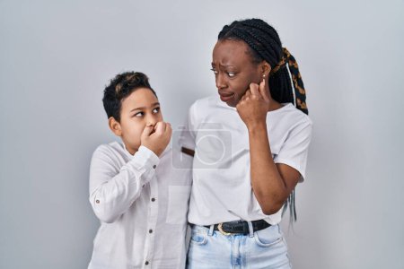 Foto de Young mother and son standing together over white background looking stressed and nervous with hands on mouth biting nails. anxiety problem. - Imagen libre de derechos