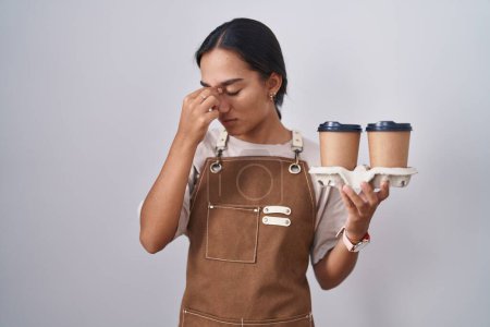 Foto de Mujer hispana joven usando delantal de camarera profesional sosteniendo café cansado frotando nariz y ojos sintiendo fatiga y dolor de cabeza. concepto de estrés y frustración. - Imagen libre de derechos