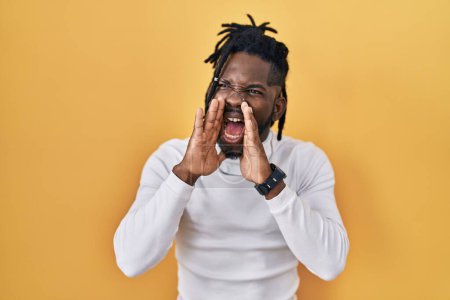 Foto de Hombre africano con rastas con jersey de cuello alto sobre fondo amarillo gritando furioso con las manos sobre la boca - Imagen libre de derechos