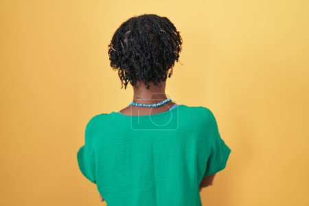 Foto de Mujer africana con rastas de pie sobre fondo amarillo de pie hacia atrás mirando hacia otro lado con los brazos cruzados - Imagen libre de derechos