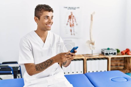 Téléchargez les photos : Jeune homme hispanique travaillant comme physiothérapeute à l'aide d'un smartphone dans une salle de physiothérapie - en image libre de droit